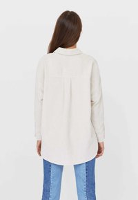 Light beige corduroy shirt with a relaxed fit, long sleeves, and a collar. Features a back pleat and rounded hem. Paired with blue jeans.