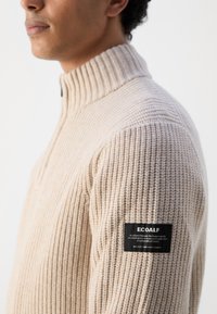 Suéter de punto acanalado beige con cuello alto, presenta un parche del logo Ecoalf en negro en el hombro izquierdo, fabricado en tejido de punto texturizado.