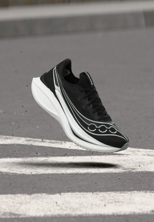 Fekete-fehér Saucony futócipő lebeg a szürke aszfalt felett, fehér útjelzésekkel, szennyeződésrészecskék lebegnek a levegőben.