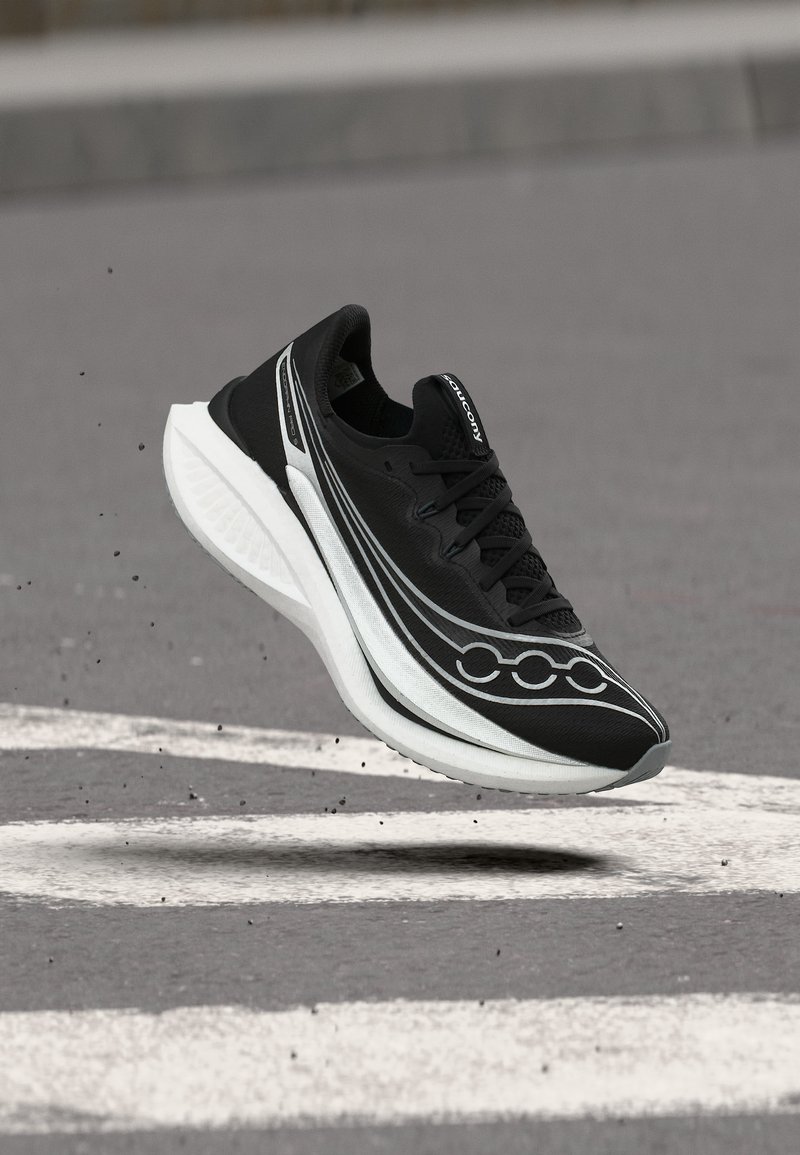 Μαύρο και άσπρο παπούτσι τρεξίματος Saucony που αιωρείται πάνω από γκρίζο πεζοδρόμιο με λευκές οδικές σημάνσεις, σωματίδια βρωμιάς αιωρούνται στον αέρα.