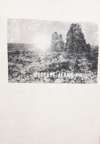 Sun shining over rugged rock formations in a desert landscape with the text "MUSTANG JEANS" at the bottom.