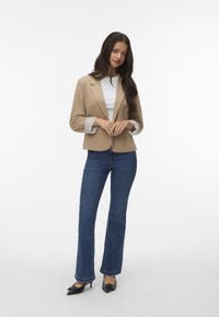 Beige blazer met een gestructureerde kraag, grijs gestreepte manchetten, gecombineerd met blauwe flared jeans en zwarte puntige hakken.