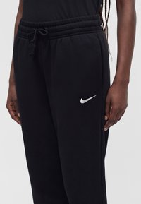 Nahaufnahme einer Person, die schwarze Nike-Sweathosen mit Kordelzugbund und weißem Nike-Swoosh-Logo am linken Oberschenkel trägt.