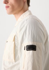 Könnyű, krémszínű dzseki gyűrött textúrával, cipzáros zsebbel és fekete anyagú felvarrón a "Barbour International" logóval.