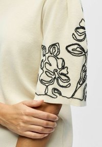 Personne tenant son bras plié portant un haut à manches courtes crème avec une broderie florale noire sur la manche.