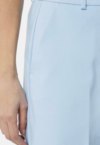 Lichtblauwe stof met een gladde textuur, met een zijnaad en een gedeeltelijke hand zichtbaar, die het stijlvolle ontwerp van het kledingstuk benadrukt.