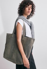 Olivgrön kordflamsväska med två långa handtag, med en strukturerad form och texturerad yta, i kombination med en ljusgrå stickad scarf.