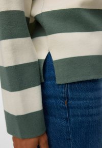 Manica e spacco laterale di un maglione a righe verdi e bianche indossato su jeans di denim blu, con una mano visibile accanto ai fianchi.