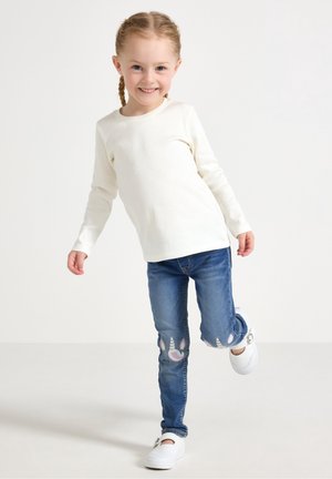 Jeune fille souriante aux cheveux tressés, portant un haut à manches longues blanc, un jean bleu avec des patchs licorne et des chaussures blanches, posant sur une jambe.