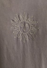 Grå stof med detaljeret hvid broderi af et solmotiv, der har et rundt ansigt og strålende solstråler. Glat tekstur, detaljeret syning.