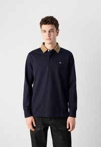 Polo a maniche lunghe blu navy con colletto in velluto a coste beige, caratterizzato da un piccolo logo sul petto, abbinato a pantaloni di jeans scuri.