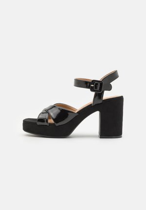 Sandalias de tacón de charol negro con un grueso tacón de bloque de ante y diseño de tira cruzada; correa en el tobillo ajustable con un hebilla metálica.