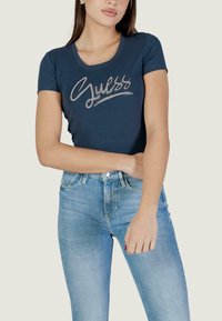 T-shirt aderente blu scuro con maniche corte, caratterizzata da un logo "Guess" argentato decorato con strass, abbinata a jeans azzurri.