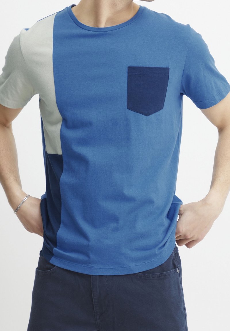 Homme portant un T-shirt à manches courtes, bicolore bleu et gris avec une poche poitrine bleu foncé, et un short foncé, les mains posées sur les hanches.