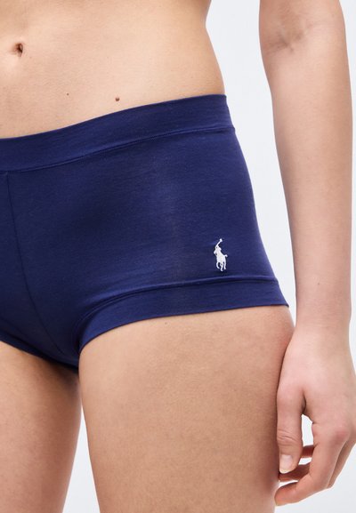 Κοντινή λήψη ενός ατόμου που φοράει σκούρο μπλε στενά σορτς με ένα μικρό λευκό λογότυπο παίκτη πόλο στο αριστερό ισχίο.