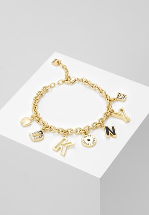 Zlatý řetízkový náramek s přívěsky včetně srdce, písmen D, K, kulaté destičky s nápisem „DKNY Circa 1989“, písmen N a Y, na bílém povrchu.