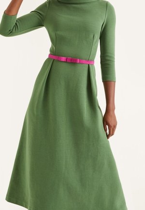 Kobieta w zielonej sukience z długim rękawem, z dopasowaną talią i cienkim różowym paskiem z kokardką z przodu.