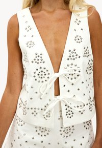 Mujer vestida con un conjunto de chaleco sin mangas y pantalones cortos blancos, adornado con tachuelas cuadradas plateadas en patrones florales, sujeto con dos lazos delanteros.