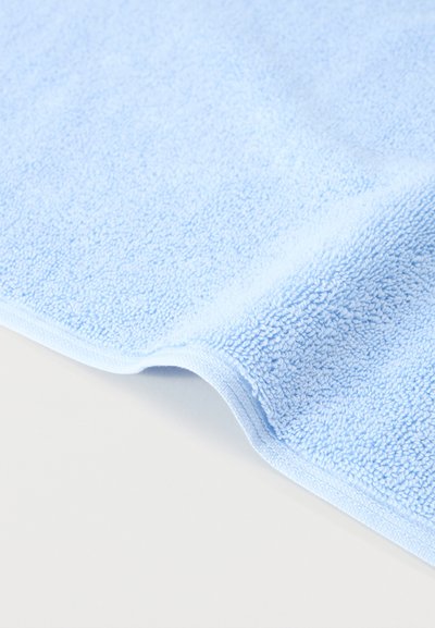 Απαλή πετσέτα από περκάλι σε ανοιχτό γαλάζιο, διπλωμένη με εμφανείς υφασμάτινες θηλιές, σε απλή λευκή επιφάνεια.