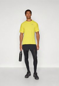 Kollane lühikeste varrukatega sportlik T-särk, mustad retuusid ja mustad spordijalatsid. Hoidmas musta veepudelit. Lihtne, funktsionaalne disain.