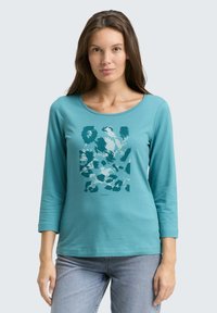 Eine Frau trägt ein türkisfarbenes Langarmshirt mit einem dunkel-türkisfarbenen abstrakten Muster und eine helle Jeans, während sie vor einem schlichten hellen Hintergrund steht.