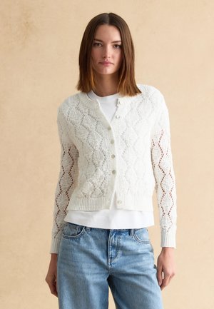 Jeune femme aux cheveux bruns et raides, portant un cardigan en maille blanche à motifs par-dessus une chemise blanche et un jean bleu sur un fond beige.