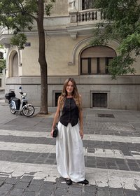 Mujer con cabello largo, vestida con top negro y pantalones blancos anchos, de pie en un paso de peatones en una calle de la ciudad cerca de un edificio de piedra beige y un scooter aparcado.