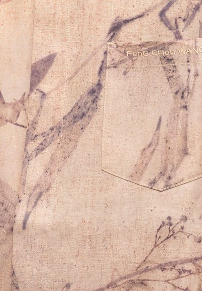 Μπεζ ύφασμα με απαλό βοτανικό σχέδιο και ραμμένη τσέπη που δείχνει μερικό χρυσό κεντημένο κείμενο "Fene Chien WANG".