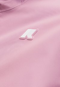 Ροζ ύφασμα με ανάγλυφο λευκό γεωμετρικό λογότυπο που παρουσιάζει ένα στυλιζαρισμένο "R" ραμμένο κοντά στην περιοχή του λαιμού.