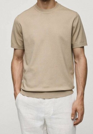 Hombre con suéter de punto beige de manga corta y pantalones de lino blancos, con las manos relajadas a los lados.