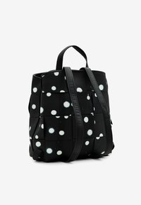 Czarny plecak z materiału w białe kropki, o kwadratowym kształcie, z dwoma uchwytami i regulowanymi paskami. Teksturowany materiał z detalami przeszyć.