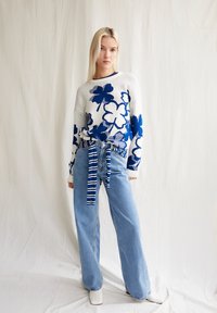 Weißer Strickpullover mit blauem Blumenmuster, kombiniert mit hellblauen, hochtaillierten Jeans. Die Jeans haben einen gestreiften Stoffgürtel. Weiße Stiefeletten.