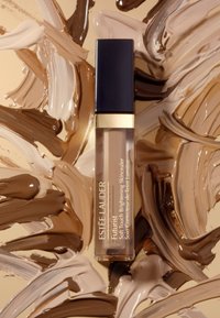 Il correttore Futurist di Estee Lauder in un tubo trasparente con accenti dorati e un cappuccio blu navy, posato su sfumature di crema marrone e beige swirlate.