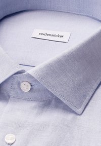 Camicia azzurra chiara con colletto rigido, chiusura a bottoni e texture tessuta. L'etichetta recita "seidensticker" cucita all'interno del colletto.