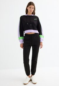 Sudadera corta negra con acentos en púrpura y verde, capucha con cordón, combinada con pantalones jogger negros con puños elásticos y zapatillas blancas.
