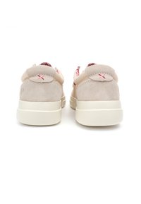 Zapatillas de ante beige con suela de goma blanca, que presentan un cuello acolchado y un interior con patrón rojo. La parte trasera tiene un acento decorativo en forma de X.