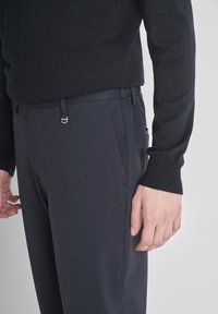 Muškarac u tamnoplavim hlačama i crnom dukserici dugih rukava, stoji uz jednoličnu svijetlu pozadinu.