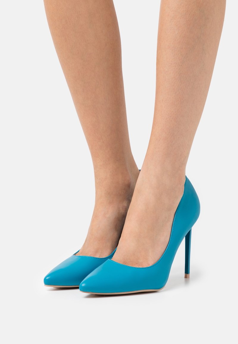 Pieds portant des escarpins bleu vif à bout pointu avec des talons aiguilles sur fond blanc.