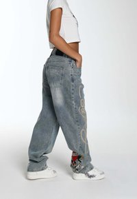 Lyseblå baggy jeans med en falmet tekstur og guld skrift i kursiv langs siden, parret med hvide sneakers med grafiske detaljer.