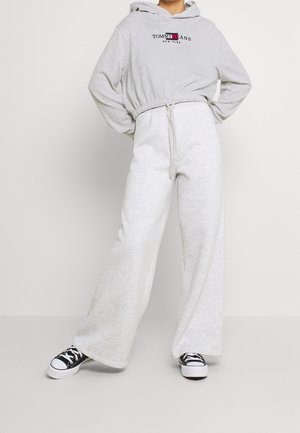Personne portant un sweat à capuche court Tommy Jeans gris clair, un pantalon large assorti et des baskets noir et blanc, debout avec une jambe légèrement en avant.