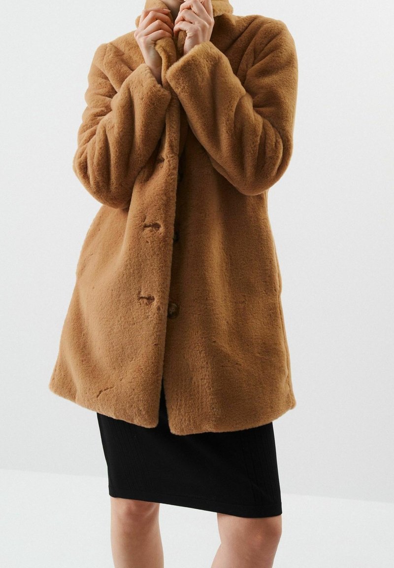 Personne portant un manteau en fausse fourrure de couleur camel mi-long, par-dessus une jupe noire arrivant aux genoux, tenant le col du manteau près de son visage.