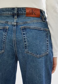 Jeans din denim de culoare albastră medie, cu talie înaltă, având două buzunare la spate și o etichetă din piele maro cu sigla Calvin Klein pe talie.