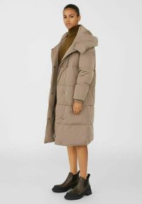 Manteau en duvet de couleur beige clair, longueur genou avec capuche, fermeture éclair pouvant être accompagnée de boutons-pression, design matelassé, associé à des bottes noires à cheville.