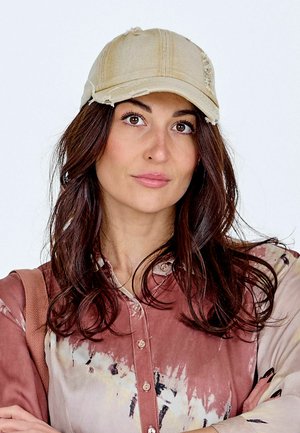Junge Frau mit langen braunen Haaren, die eine beige abgenutzte Baseballkappe und ein geknöpftes, gemustertes rosafarbenes Hemd trägt, schaut nach vorne.