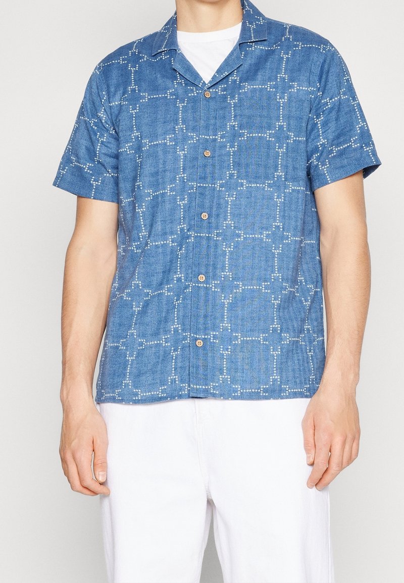 Modrá košile s krátkým rukávem z džínoviny s bílým geometrickým vzorem a hnědými knoflíky. Lehká s volným střihem.