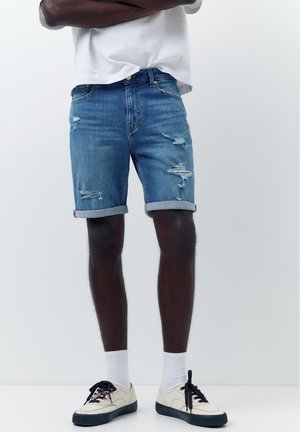 Personne portant un short en jean déchiré avec revers, des baskets blanches avec semelles et lacets noirs, des chaussettes blanches hautes et un T-shirt blanc, debout les bras croisés.