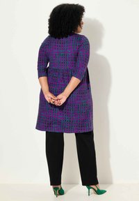 Paarse tuniek met patroon, voorzien van driekwartmouwen en een peplum taille, gedragen over zwarte broeken, gecombineerd met groene hoge hakken.