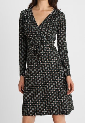Kvinde iført en sort slå-om-kjole med lange ærmer og et lille blomsterprint i hvidt, grønt og rødt, bundet i taljen.