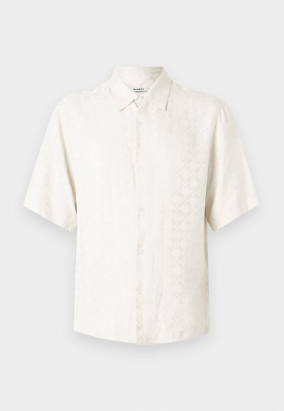 Λευκό πουκάμισο με κοντό μανίκι και κουμπιά, με διακριτικό υφασμάτινο σχέδιο και μυτερό γιακά, σε απλό φωτεινό φόντο.