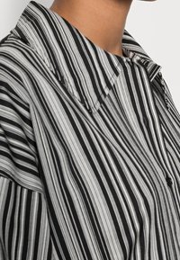 Chemise rayée noire et blanche avec un col pointu, une patte de boutonnage à l'avant, et une coupe ample. Le tissu semble lisse avec des lignes verticales étroites.